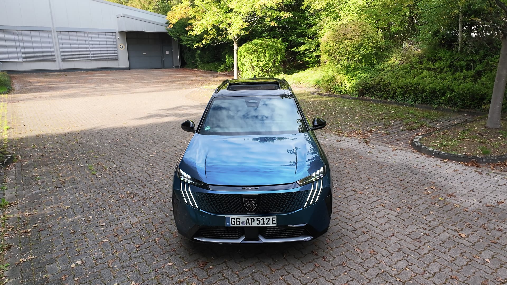 Blauer Peugeot 5008 von vorn und leicht von oben auf einem gepflasterten Platz, dahinter Bäume, Büsche und eine Halle.