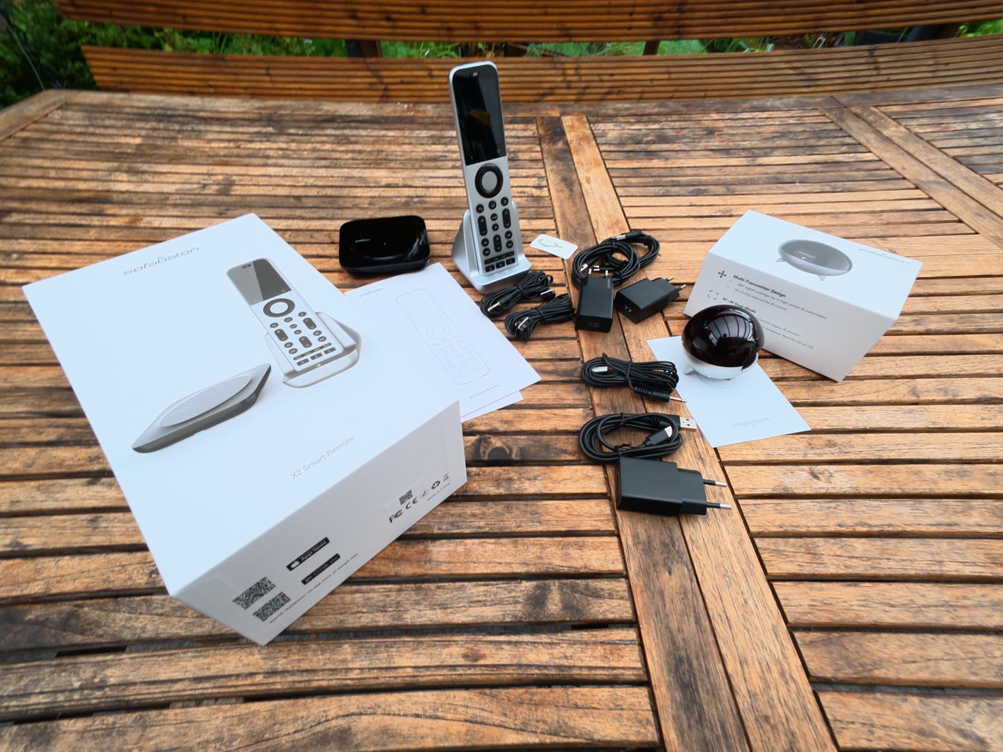 Auf einem Holztisch im Freien liegen der geöffnete Karton der SofaBaton X2 Smart Remote, die Fernbedienung in ihrer Ladestation, eine schwarze Basisstation, ein zweiter Karton mit einer runden Einheit, mehrere Netzteile, Kabel und Anleitungen.