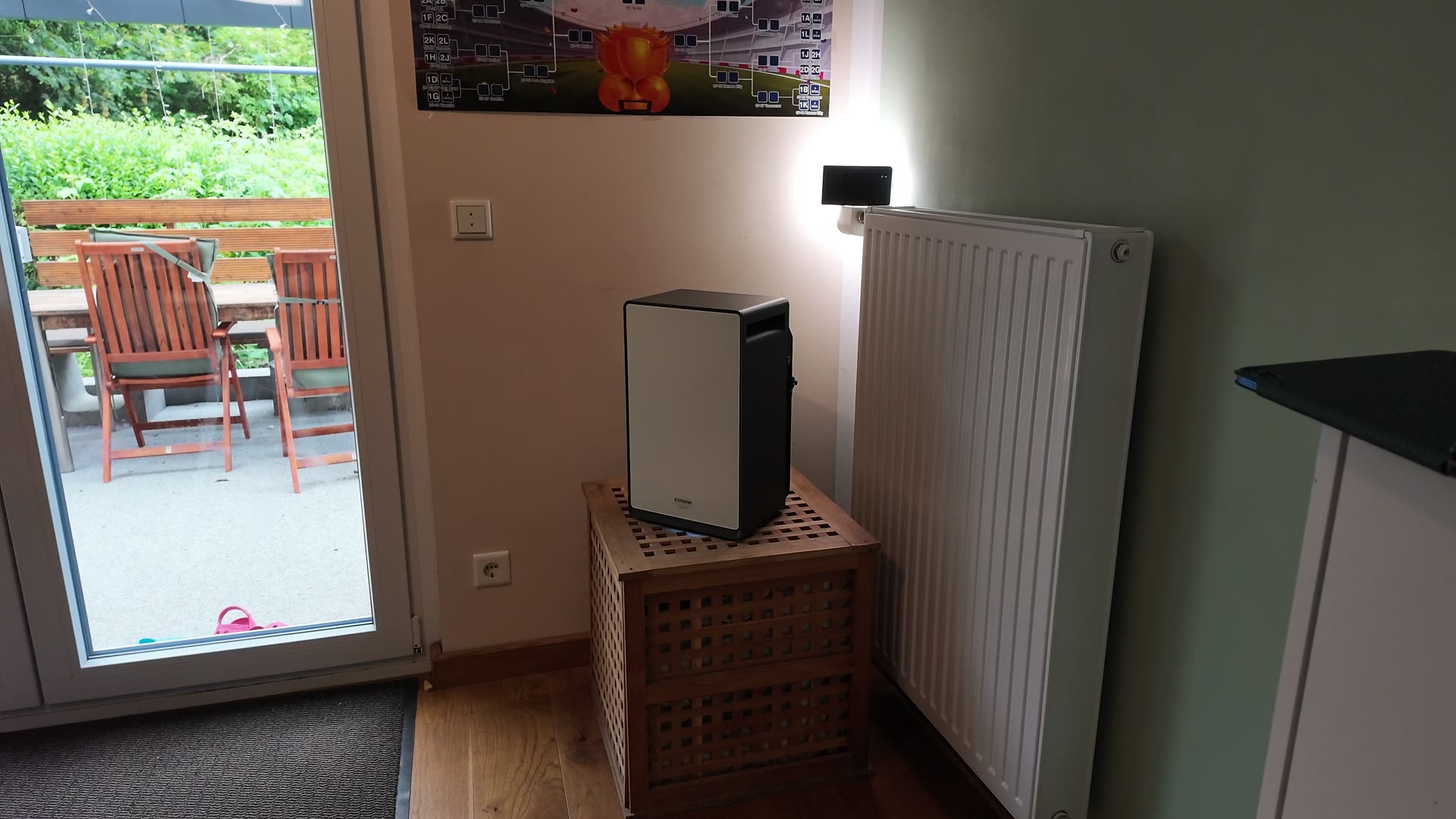 Der Speicher mit heller Front und dunklem Gehäuse steht auf einem Holzwürfel mit Gitterstruktur neben einem Heizkörper im Wohnzimmer.
