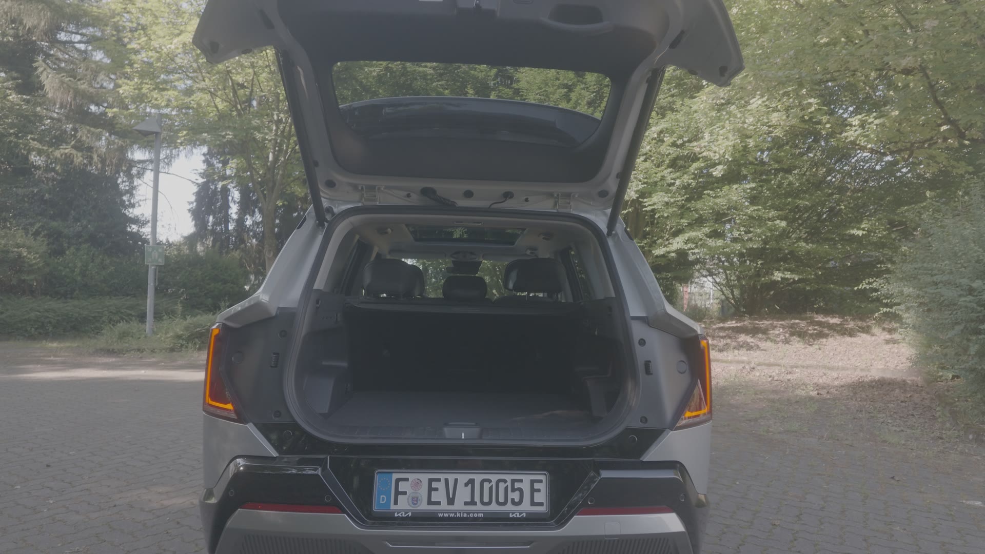 Heckansicht eines silbernen Kia mit weit geöffneter Heckklappe und leerem Kofferraum, im Hintergrund Bäume