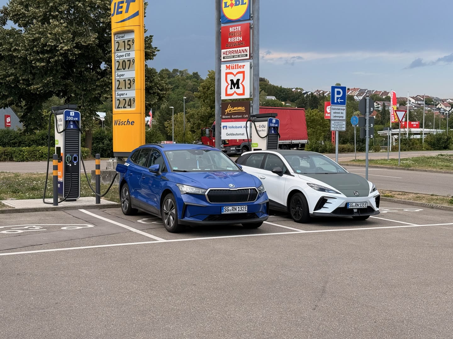 Ein blaues und ein weißes Elektroauto stehen nebeneinander auf Parkplätzen vor zwei Schnellladesäulen auf einem Parkplatz mit Tankstellen- und Marktschildern im Hintergrund.