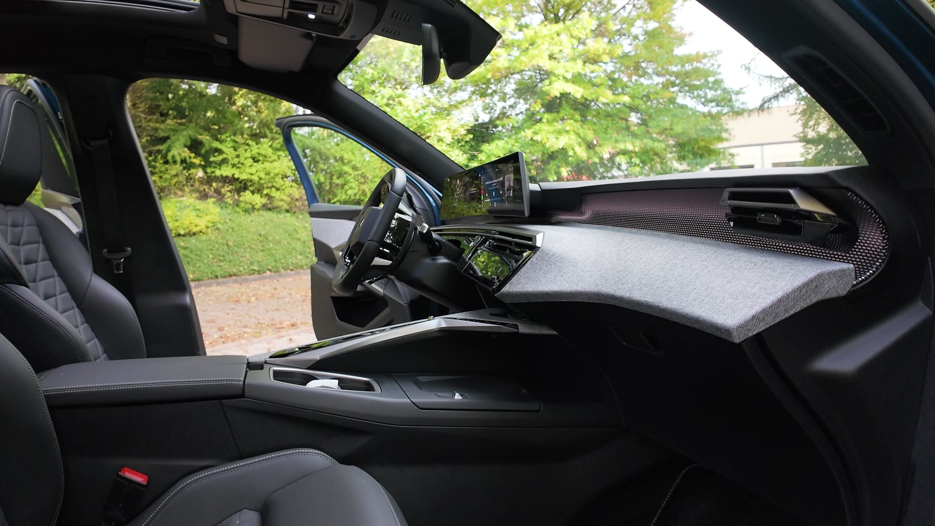 Blick in den Innenraum eines Autos: dunkles Armaturenbrett mit Stoffoberfläche, Lenkrad, mehrere Bildschirme und schwarze Ledersitze, die Fahrertür steht offen