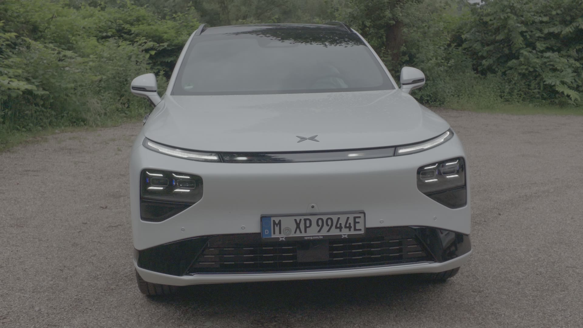 Frontansicht eines weißen XPENG-Elektro-SUV auf einem Kiesplatz vor Büschen und Bäumen
