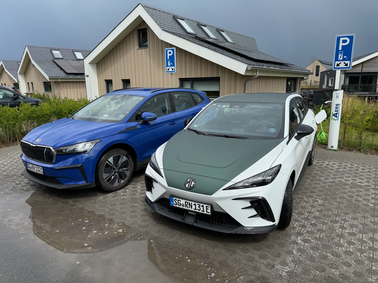 Ein blaues SUV und ein weißes Auto mit grüner Motorhaube und MG-Logo stehen nebeneinander auf Pflastersteinen, daneben eine Ladesäule mit Parkschild und Häuser mit Holzfassade im Hintergrund.