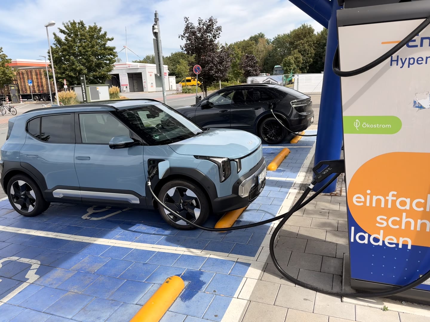 Hellblaues Elektroauto steht an einer Schnellladesäule auf einem blau markierten Ladeplatz und ist mit dem Ladekabel verbunden. Dahinter lädt ein dunkles Fahrzeug an der Säule.