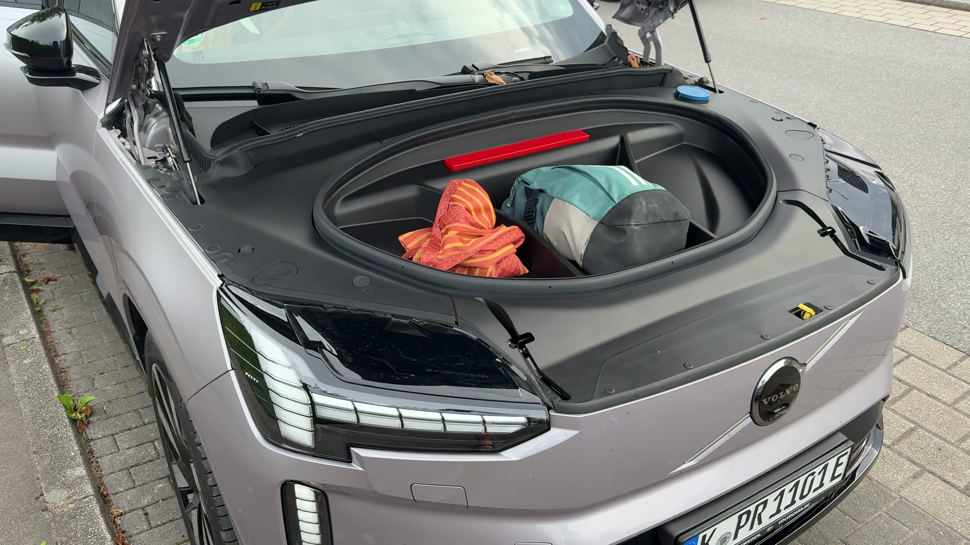 Geöffneter Frontkofferraum eines silbergrauen Volvo mit einem orangefarben gestreiften Tuch und einer Tasche