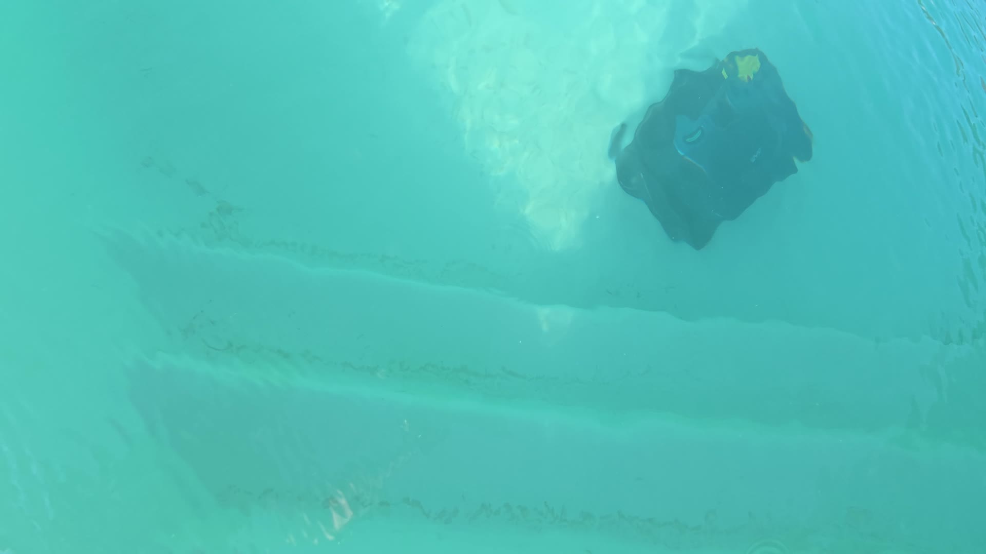 Blick von oben ins türkisfarbene Wasser: Ein dunkler Poolroboter mit gelbem Fleck bewegt sich im rechten oberen Bildteil über den Beckenboden, im Hintergrund sind Stufen oder Absätze zu erkennen.
